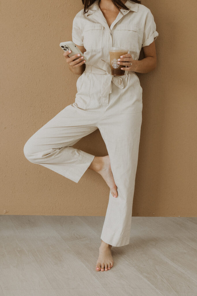 A woman standing against a wall, checking her iPhone and holding a Starbucks iced coffee.